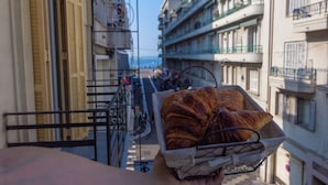 Un balcon avec un panier de croissants, donnant sur une rue bordée d’immeubles et offrant une vue sur la mer.