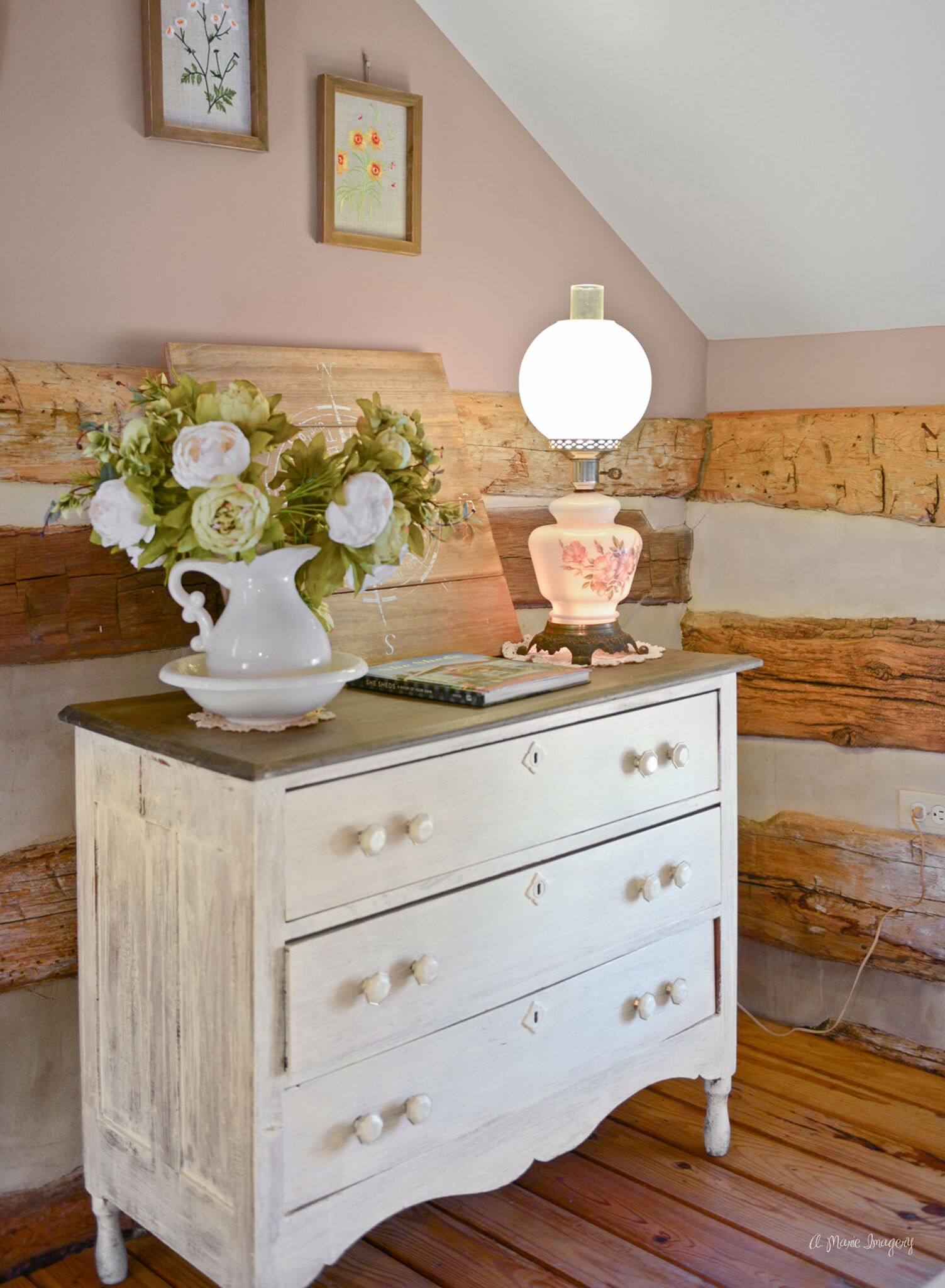 A white dresser with three drawers, a vase of flowers, and a lamp on top.