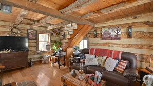 Ein rustikales Ferienhaus-Interieur mit Holzbalkendecke, einem Ledersofa mit dekorativen Kissen in Landesfarben, einem Holztisch und einem Fernseher auf einer Holzkommode.
