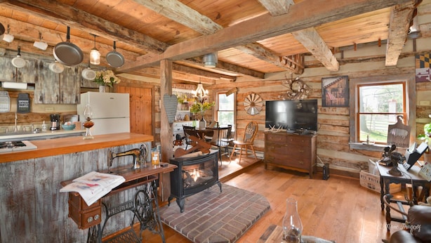 Ein rustikales Blockhaus-Interieur mit Holzbalkendecke, Kamin, Essbereich und Küche mit Kühlschrank und hängenden Töpfen.