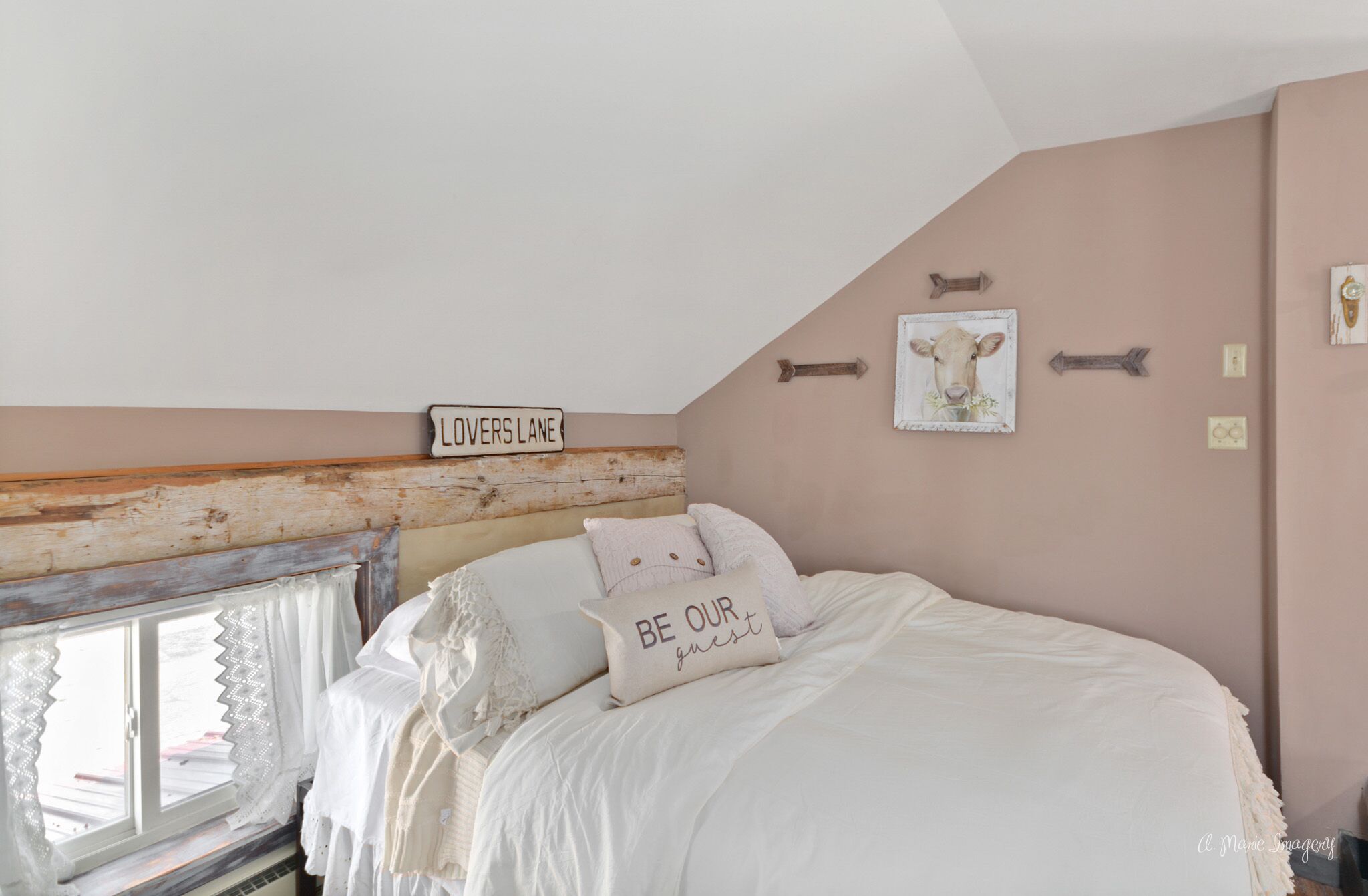 Een slaapkamer met een schuine balkenlaag, een bed met wit beddengoed, een bordje met de tekst 'Lovers Lane' en een schilderij van een koe aan de muur.