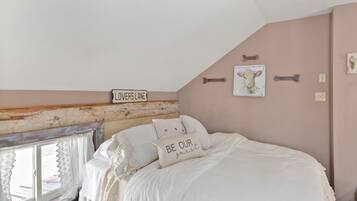 Een slaapkamer met een schuine balkenlaag, een bed met wit beddengoed, een bordje met de tekst 'Lovers Lane' en een schilderij van een koe aan de muur.