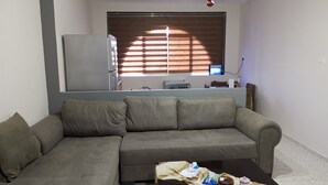 A living room with a sofa, a coffee table with items, a kitchen area with appliances, and a window with blinds. - Visit Jerusalem & Bethlehem, Quite, Safe and free Secured parking.  (Jerusalem)