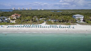 Ein Luftbild eines Strandes mit Reihen von Sonnenschirmen und Liegestühlen, einige Gebäude im Hintergrund und ein freier Blick auf das Meer.