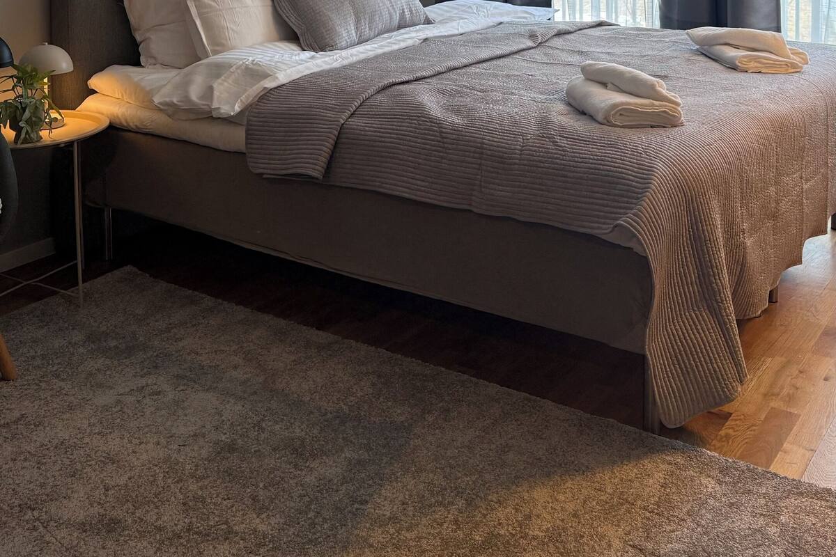 Un dormitorio con una cama grande, paredes grises, dos cuadros abstractos, una ventana con cortinas oscuras y una mesita de noche con una lámpara.