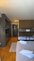 A modern hotel room with a kitchenette, a bed with two bunk beds, a wooden floor, and a door leading to the hallway.