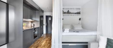 A compact room with a bed, kitchenette, and a view of a forest through a window.