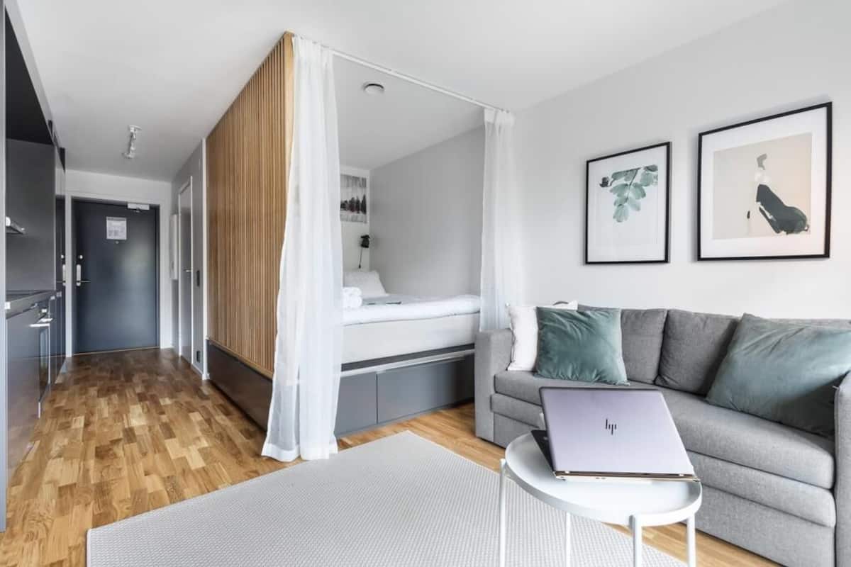 Una habitación de hotel moderna con una cama, un sofá gris, un ordenador portátil sobre una mesita y dos cuadros en la pared.