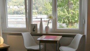 A room with a table and two chairs by a window with a view of a body of water and greenery. - Apartments Rheinblick-Koblenz (Neuendorf)