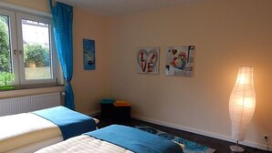 Un dormitorio con dos camas, una ventana con cortinas, un radiador, una mesita con una silla y una obra de arte en la pared.