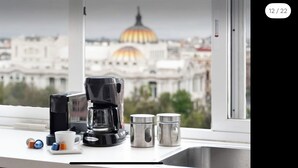 Eine Küchenarbeitsplatte mit einer Kaffeemaschine, zwei Tassen und Salz- und Pfefferstreuern. Durch ein Fenster ist eine Stadtansicht mit einem markanten, kuppelartigen Gebäude zu sehen.