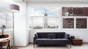Ein modernes Wohnzimmer mit einem grauen Sofa, einem Schreibtisch mit Lampe und Blick auf eine Skyline durch große Fenster.