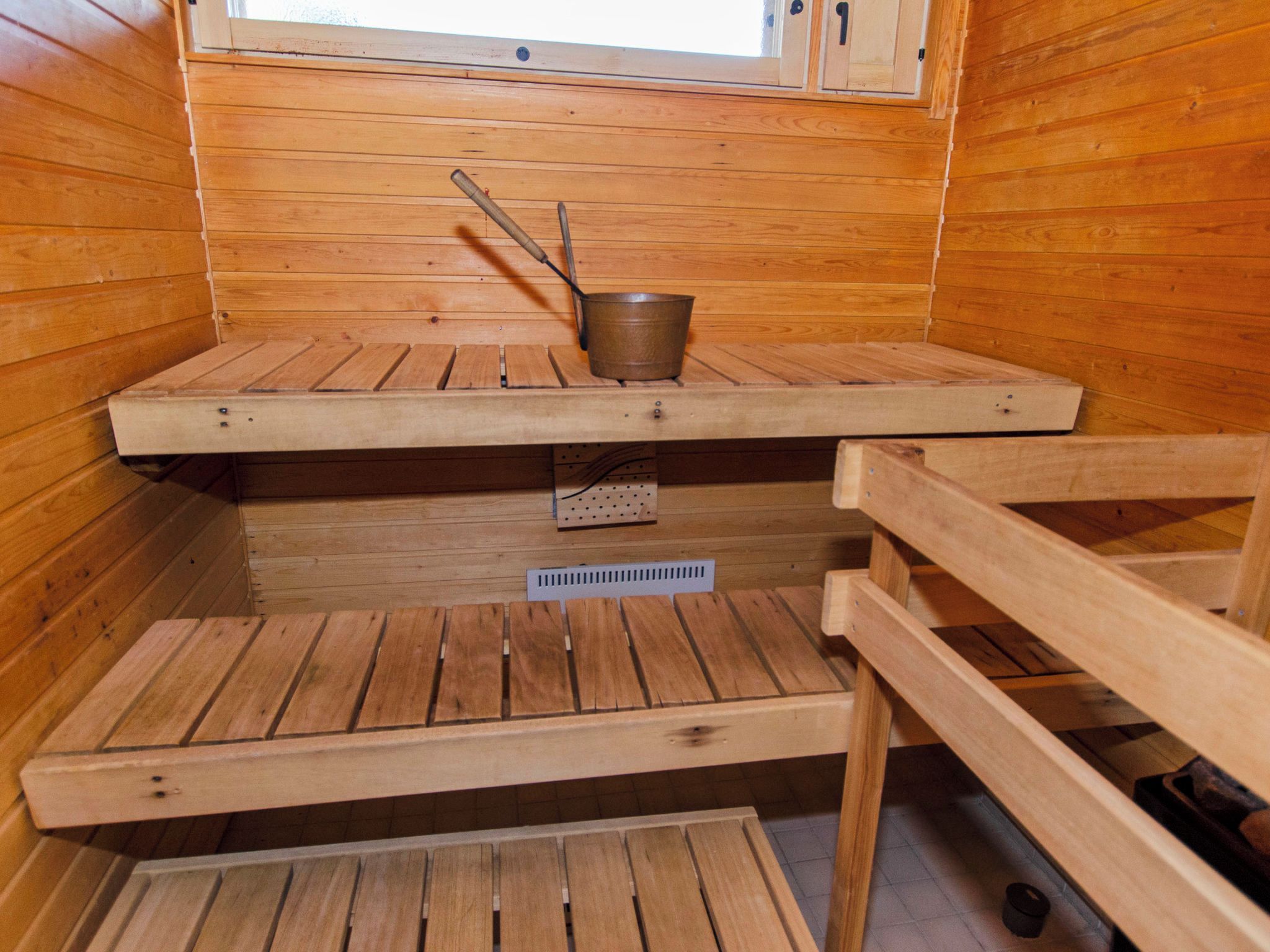 A sauna room with wooden benches, a metal bucket, and a window above.