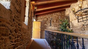 Un couloir aux murs de pierre, avec une porte en bois, une rambarde en métal et des lampes fixées au mur.
