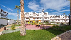 A playground with swings and slides, surrounded by palm trees and a building with balconies. - Apartamento Oregano (Mar de Pulpi) (San Juan de los terreros)