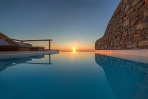 A serene infinity pool with a sunset view, stone walls, and a wooden deck with lounge chairs. - Villa Hermes (Mykonos)