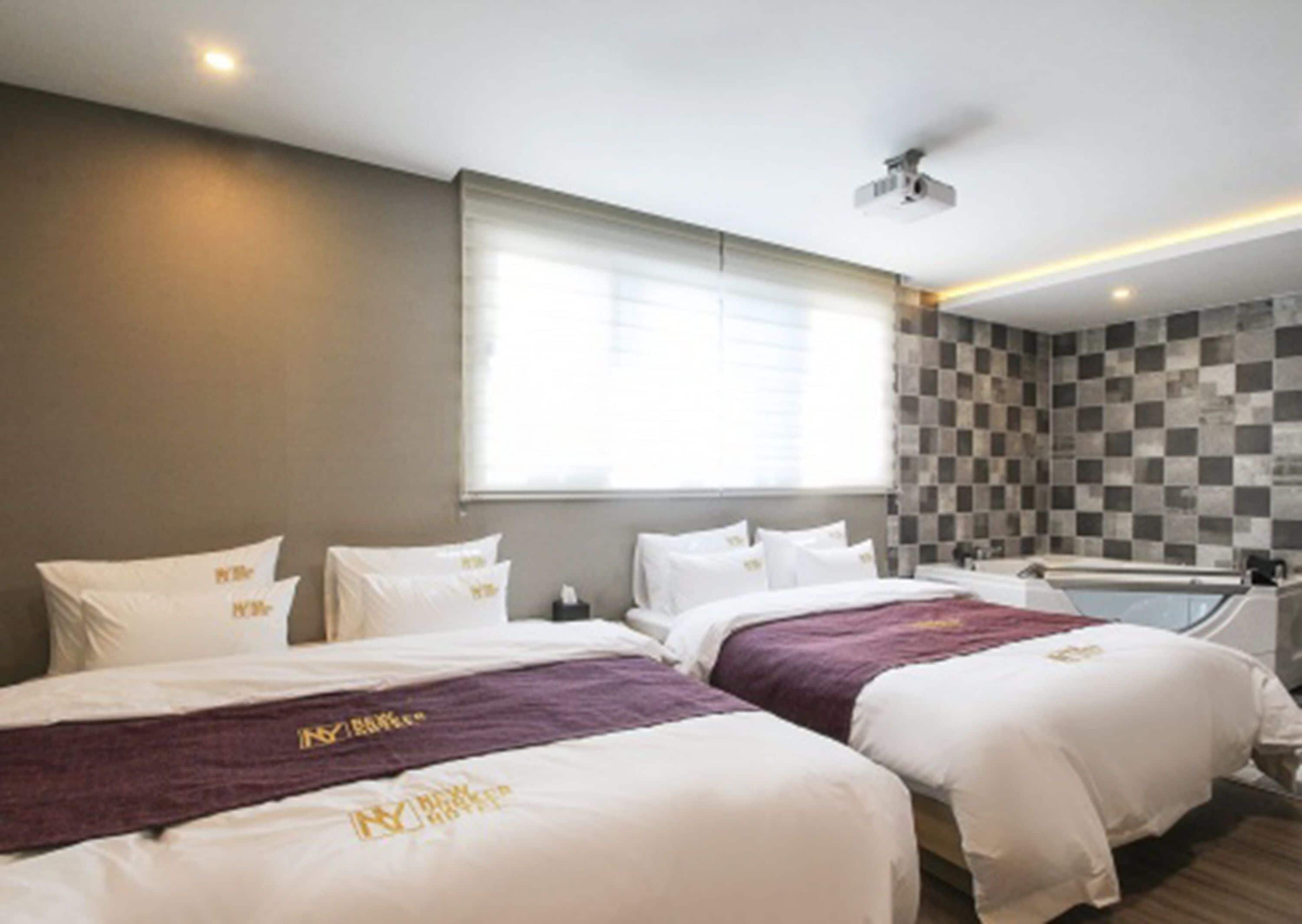 Habitación de hotel con dos camas, un ventanal amplio, un proyector en el techo y un estampado de cuadros en la pared.