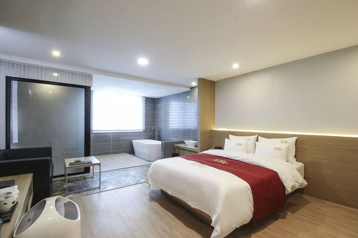 A modern hotel room with a large bed, a separate bathroom, a seating area, and a large window providing natural light.