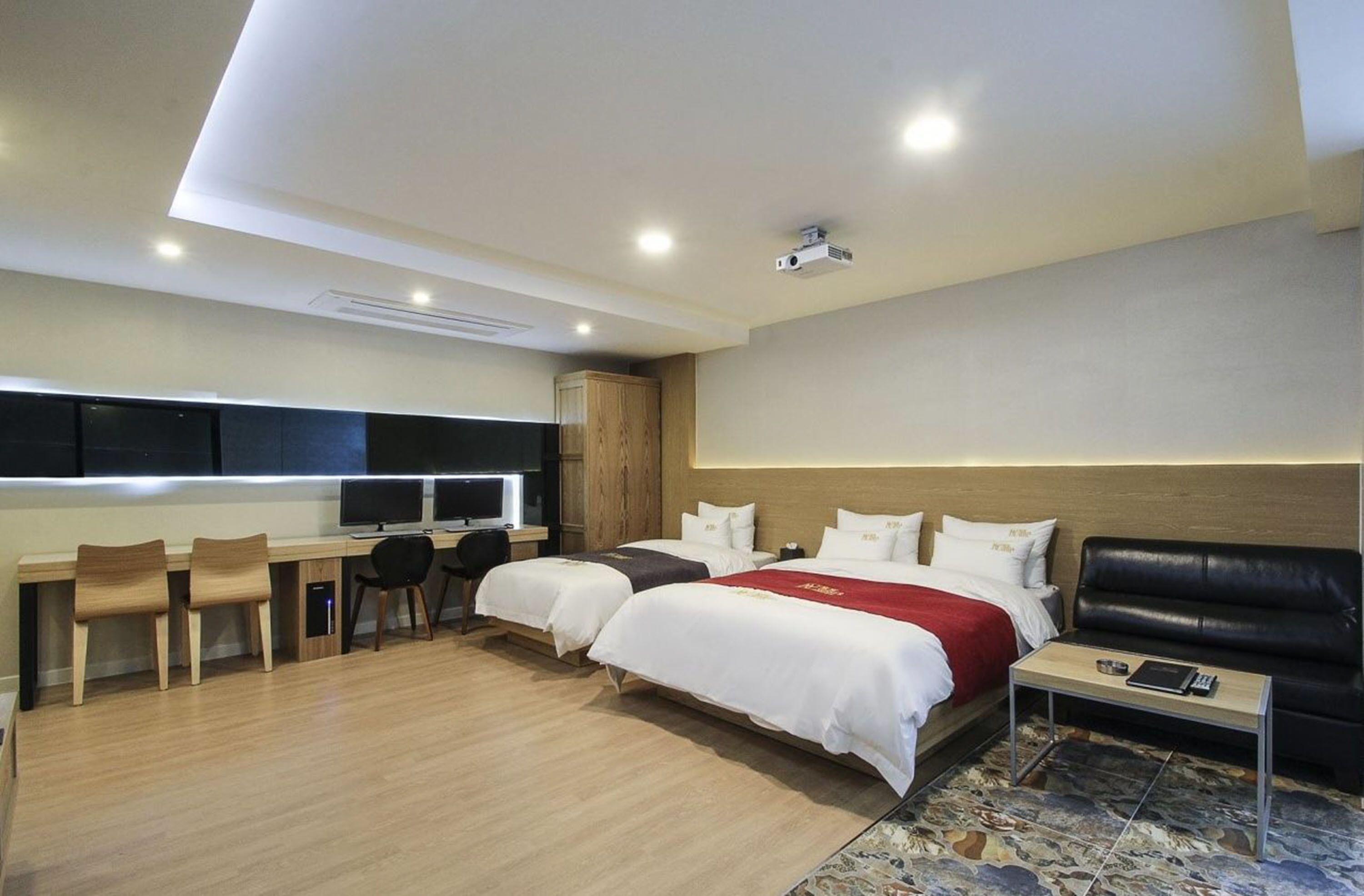 Una habitación de hotel moderna con dos camas, un escritorio con computadora, un sofá de cuero negro y una pequeña mesa con control remoto.