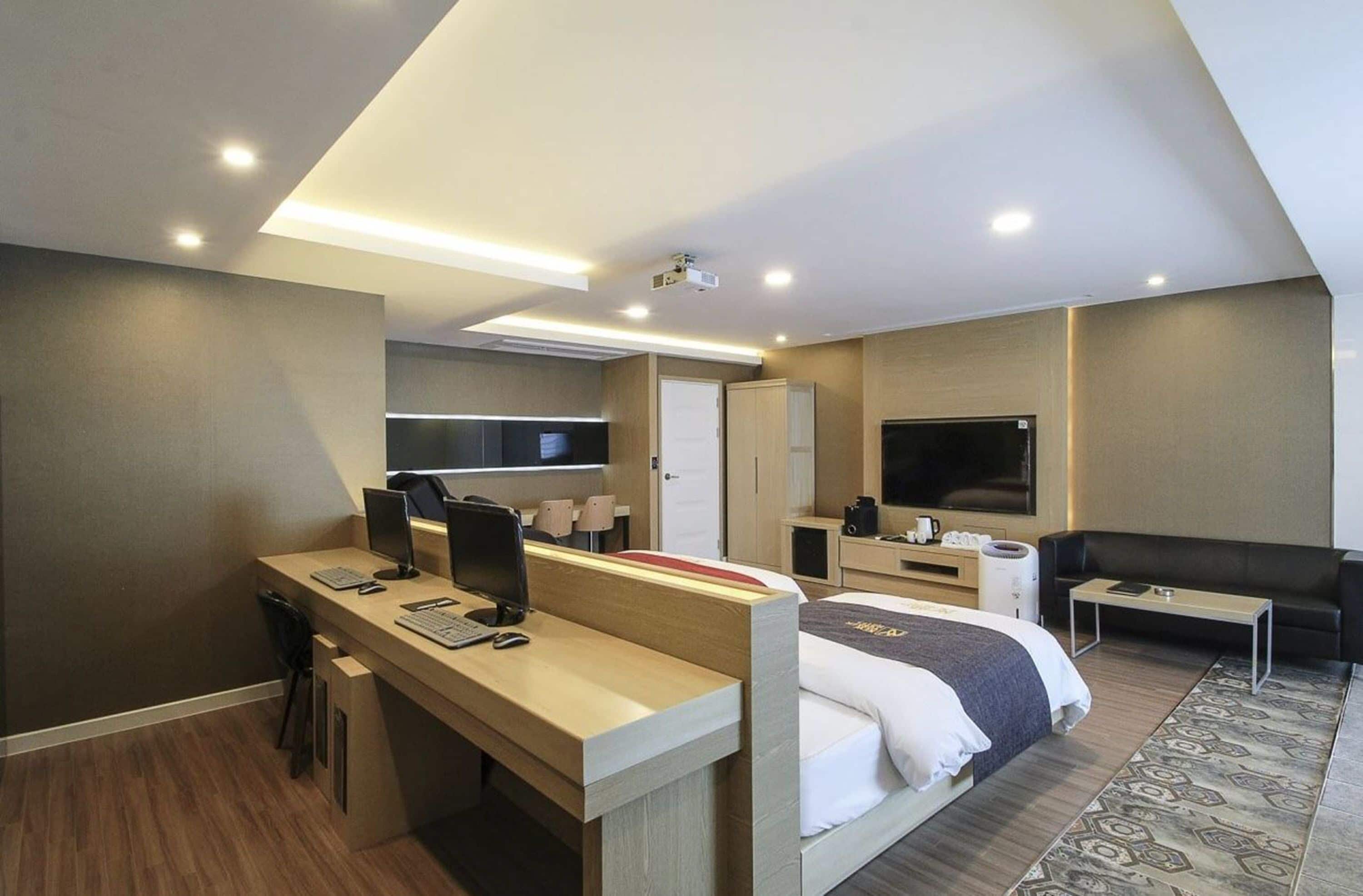A modern hotel room with a desk, computer, bed, TV, and a small seating area.