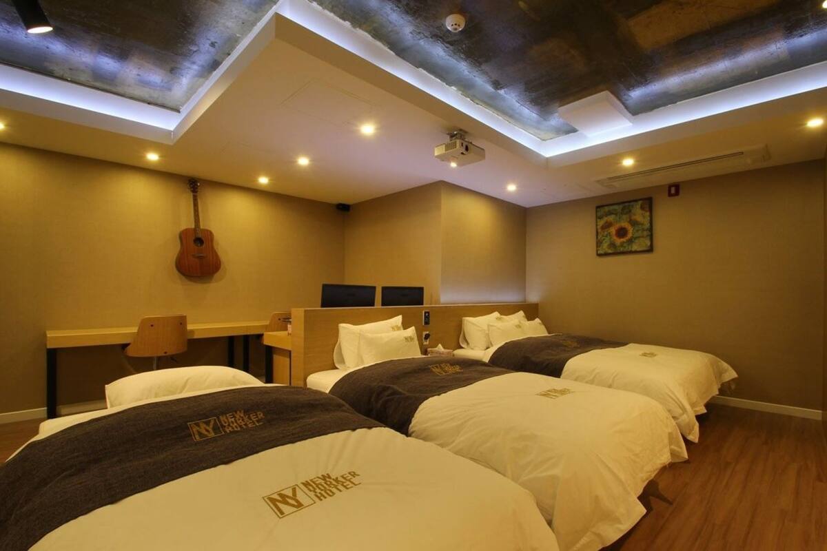 A hotel room with three beds, a guitar, a projector, and a painting on the wall.