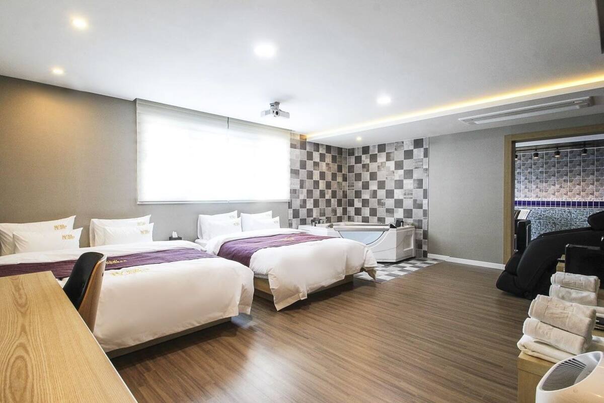 A hotel room with two beds, a bathtub, a massage chair, and a window with blinds.