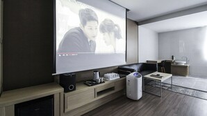 A modern living room with a large flat-screen TV mounted on the wall, a sofa, a coffee table, a small kitchen area with a sink and a microwave, and a compact entertainment unit with speakers and a gaming console. - New Yorker Hotel (Jinju)