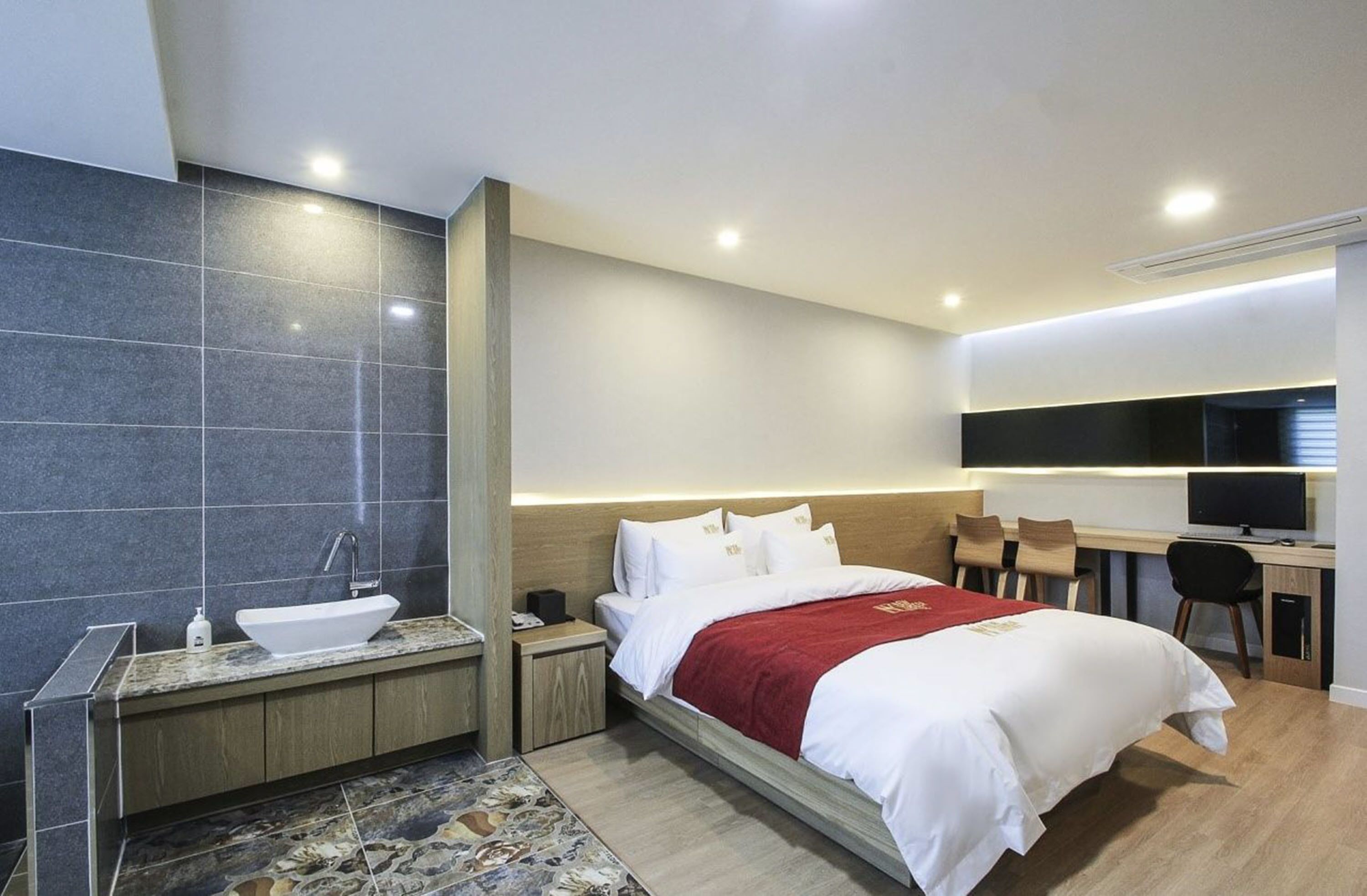 A modern hotel room with a bed, bedside table, and a desk with a chair.