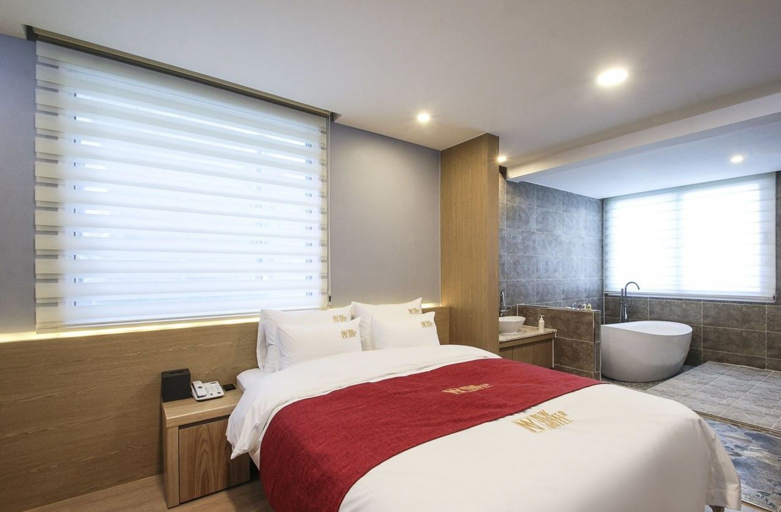 A modern hotel room with a large bed, a bedside table with a phone, a red throw blanket, a bathroom with a bathtub, and a window with blinds.