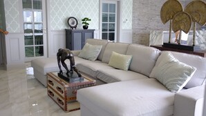 Ein modernes Wohnzimmer mit einem beigefarbenen Ecksofa, einem Glastisch und einer dekorativen Pferdestatue. Ein Schrank mit einer Uhr und einer kleinen Pflanze steht darauf. Der Raum hat einen Marmorboden und raumschallabsorbierende Tapete.