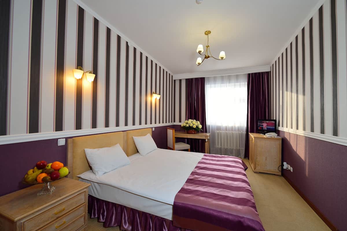 A hotel room with a large bed, a bedside table with a fruit bowl, a desk with a computer, and a TV.