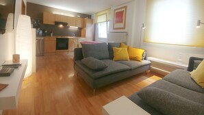 A modern living room with a grey sofa, yellow cushions, and a white coffee table. A kitchen with wooden cabinets and a dining area is visible in the background. - Apartments Rheinblick-Koblenz APARTMENT - 2 (Neuendorf)