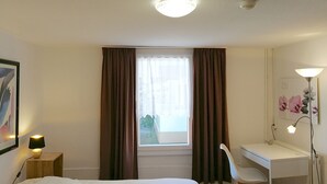 A bedroom with a bed, a desk, a chair, a lamp, and a window with curtains. - Apartments Rheinblick-Koblenz APARTMENT - 2 (Neuendorf)