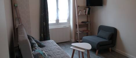 A living room with a sofa, a chair, a small table, a bookshelf, and a television mounted on the wall.