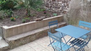 Une petite terrasse extérieure avec une table et des chaises en métal bleu, un bac à fleurs en béton planté, et un mur en pierre.