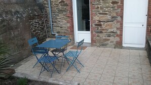 Un bâtiment en pierre avec un patio carrelé, une table entourée de chaises bleues et une porte blanche.