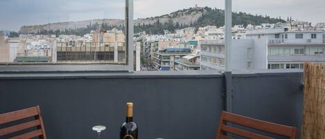 A balcony with a table, two chairs, a bottle of wine, and two wine glasses. A cityscape with buildings and a hill is visible in the background.