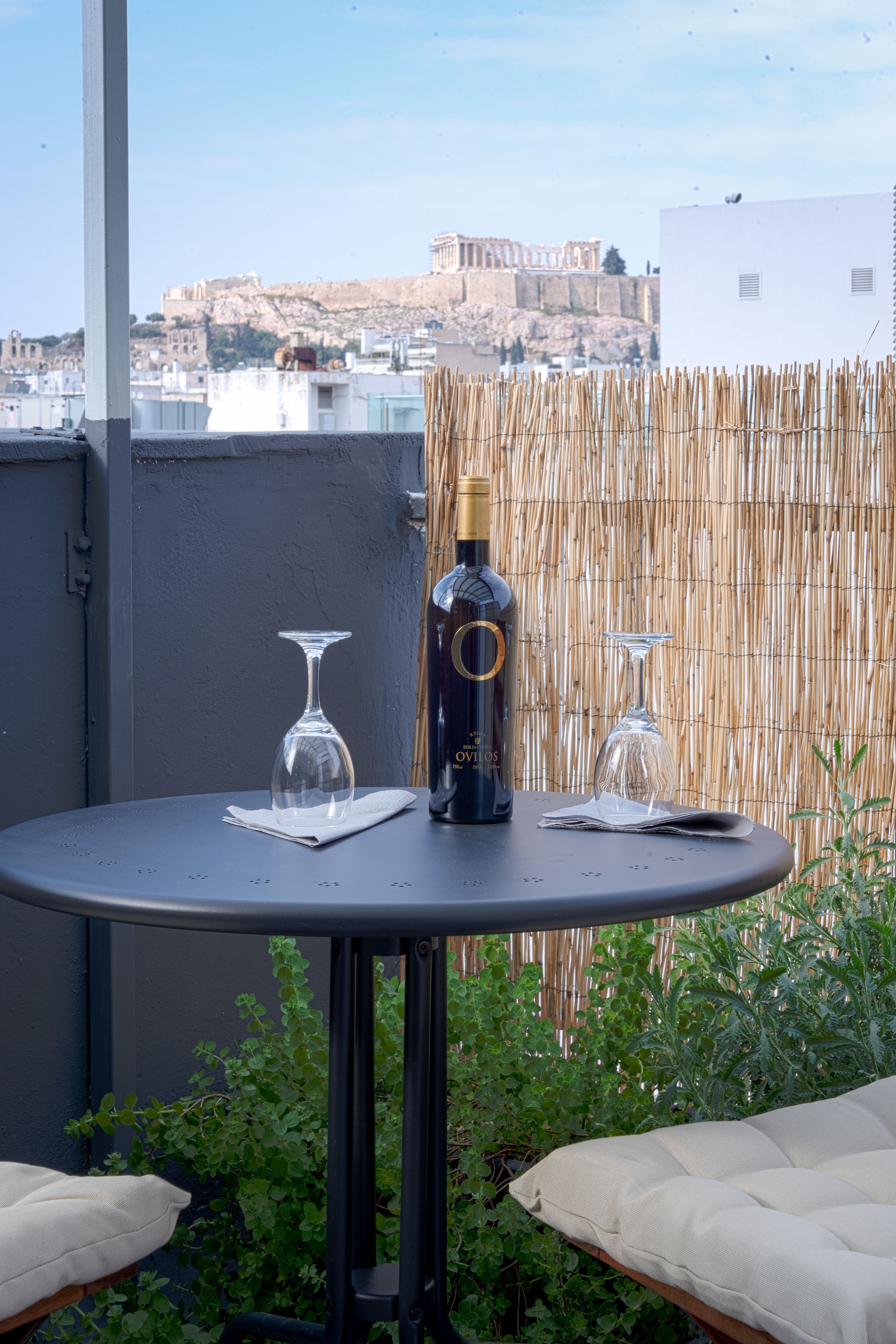 A round table with a bottle of wine and two wine glasses on a balcony with a view of ancient ruins in the background. - Nest Studio with Terrace, Acropolis View (Athens)