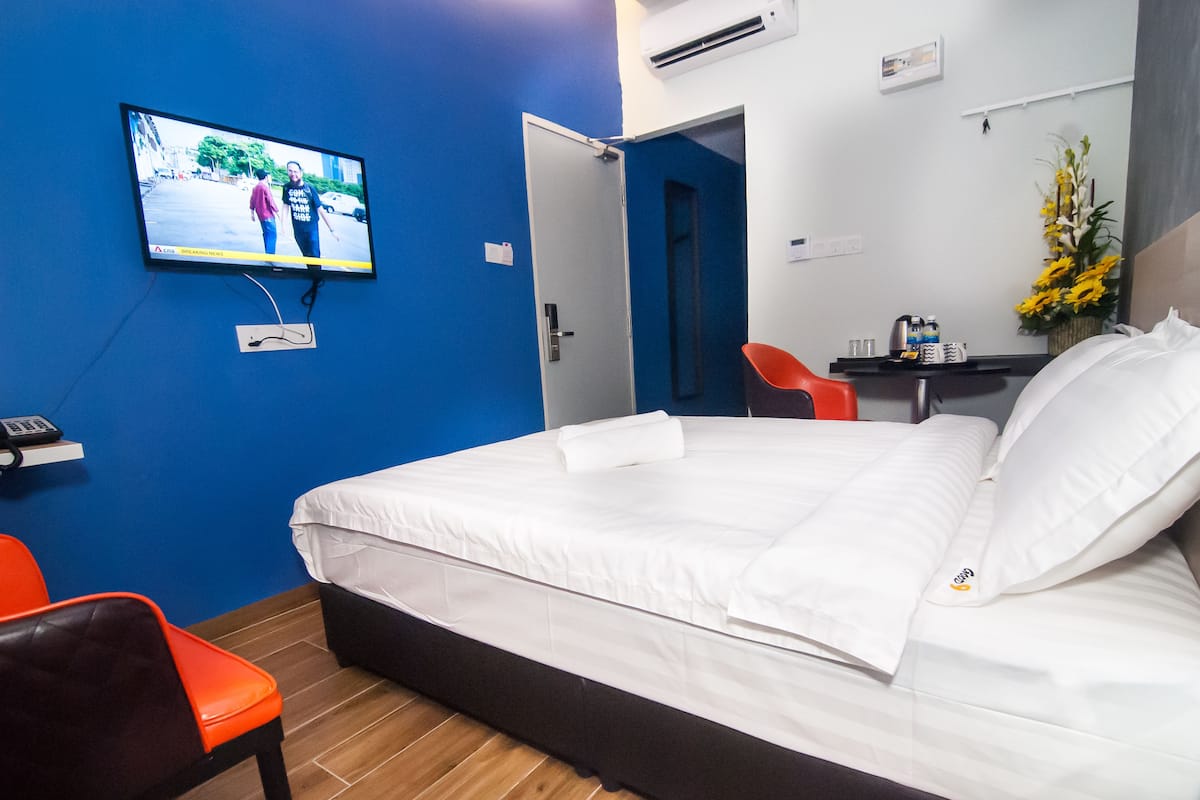 A hotel room with a bed, a TV mounted on the wall, a desk with a chair, and a small table with a vase of flowers.
