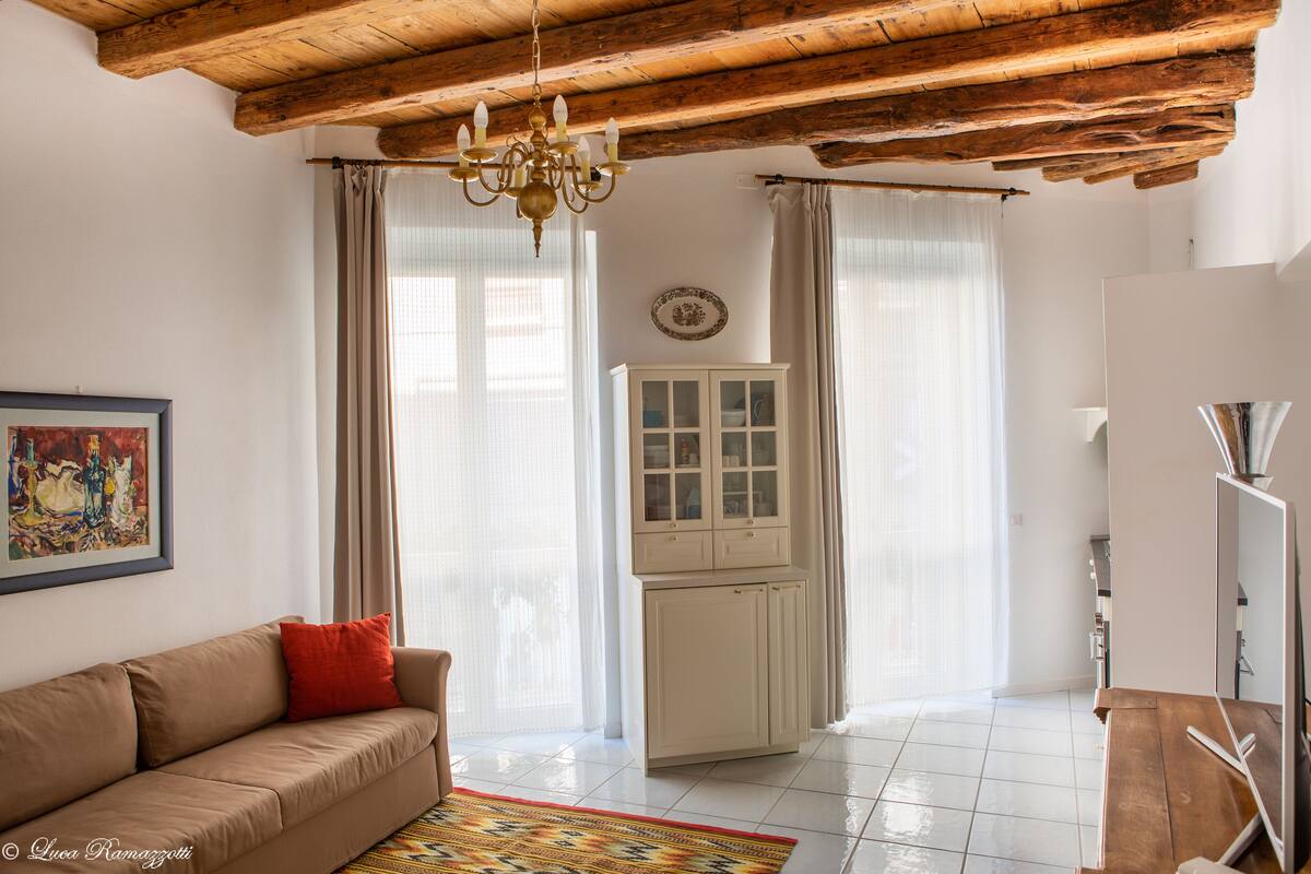 Een woonkamer met een beige bank, een houten plafond, een kroonluchter, een kast en een groot raam met gordijnen.
