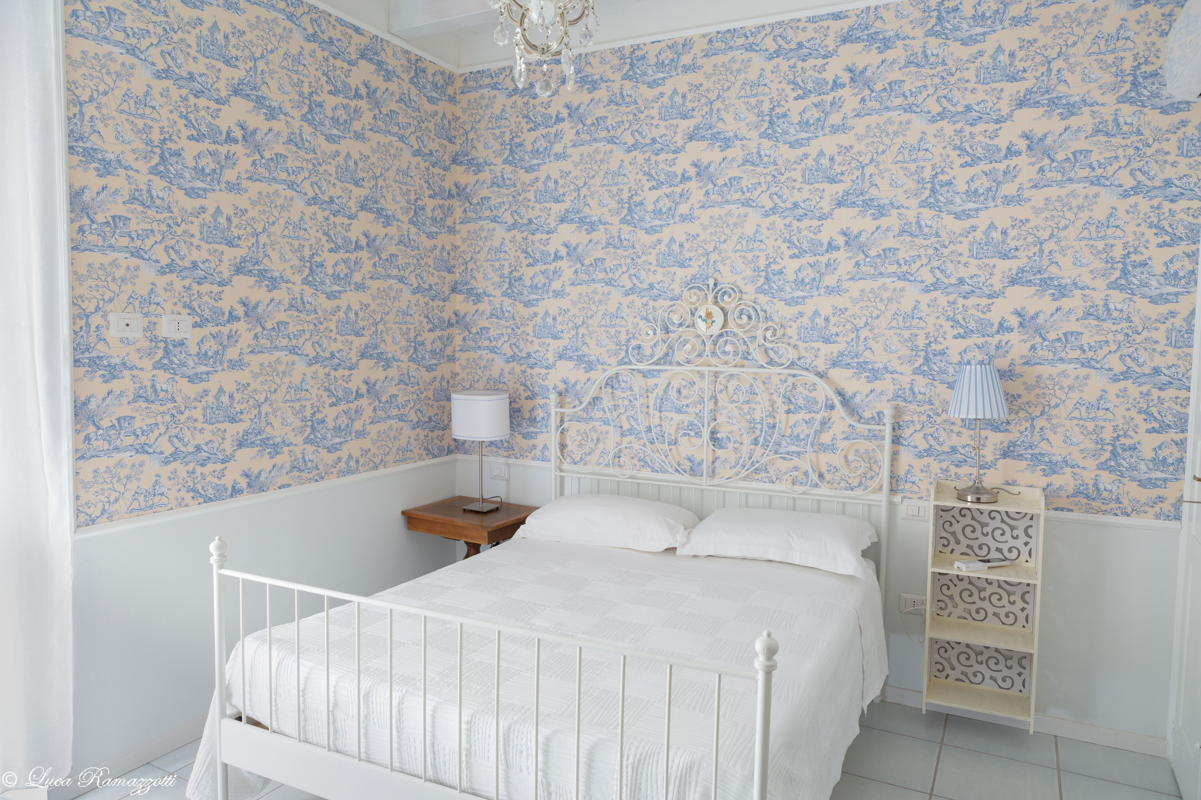 Un dormitorio con una cama blanca, una mesita de noche, una lámpara y un estante decorativo.