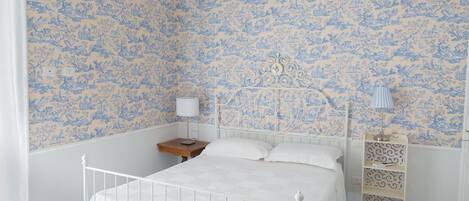 Un dormitorio con una cama blanca, una mesita de noche, una lámpara y un estante decorativo.