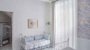 A room with a white bed, a chandelier, a window with sheer curtains, and a patterned wallpaper. - Janna 'e Mare (Cagliari)