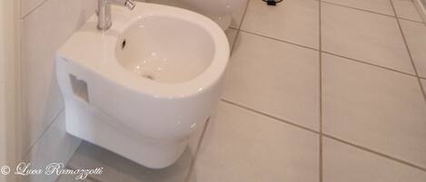 A bathroom with a bidet, toilet, and a sink. There is a framed picture on the wall and a small shelf with toiletries.