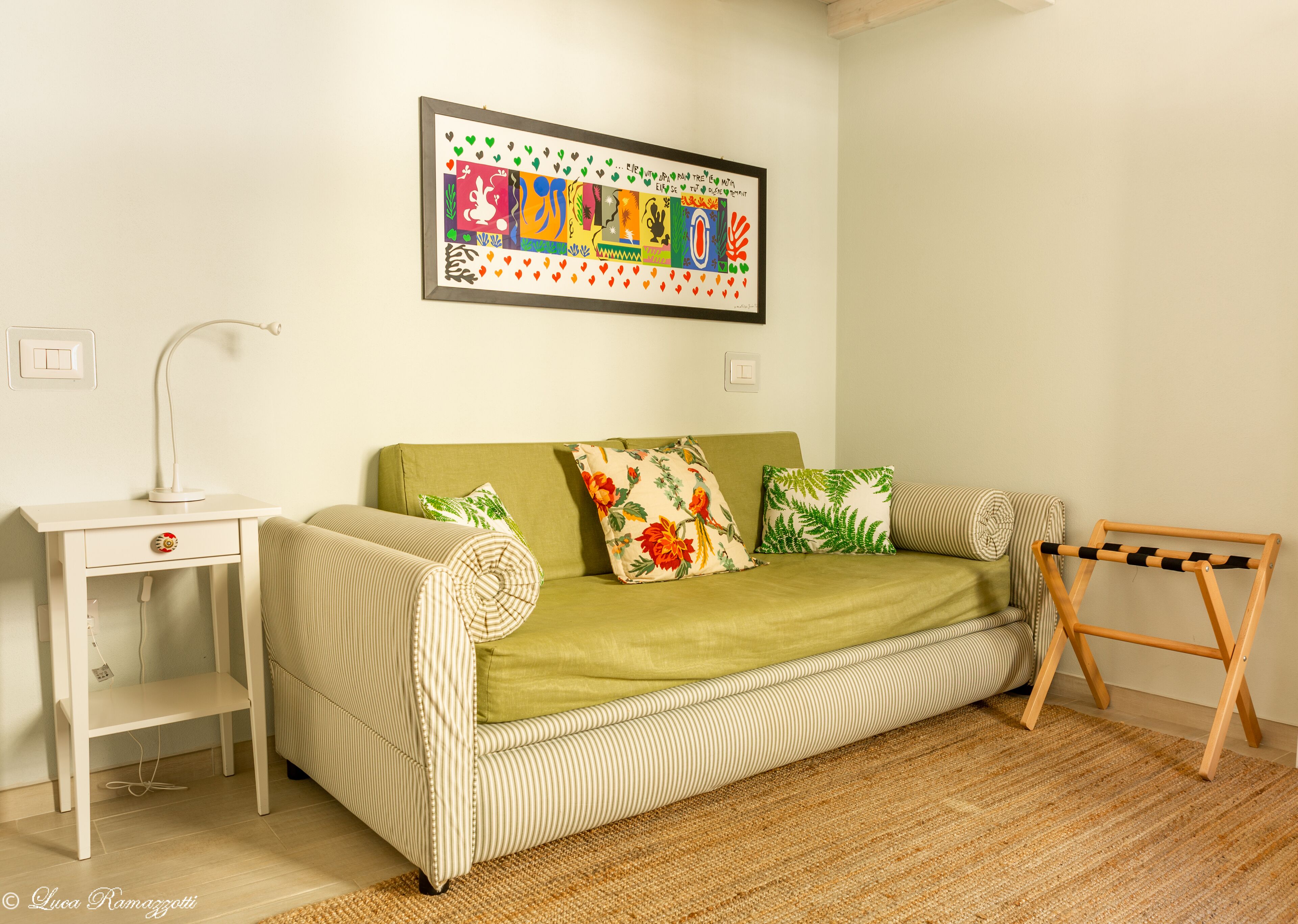 A room with a green sofa, striped pillows, a small white table with a lamp, and a framed artwork on the wall.
