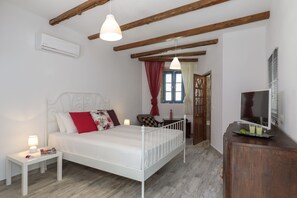 A bedroom with a white metal bed frame, a bedside table with a lamp, a TV on a wooden cabinet, and a window with red curtains. - Ai Yanni Boutique Rooms - Adults Only (Rhodes)