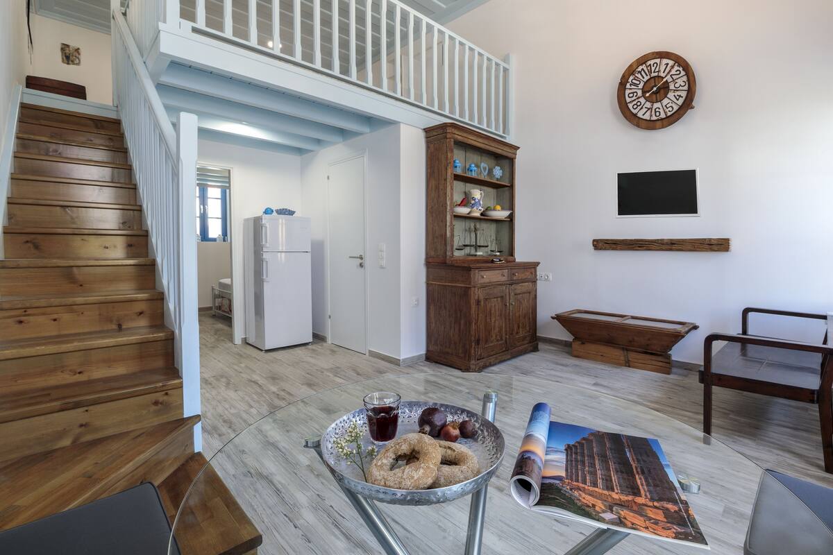 A spacious living area with a wooden staircase, a wooden cabinet, a wall clock, and a dining table.