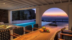 Uma área de refeições ao ar livre com mesa e cadeiras de madeira, uma cesta de frutas e vista para o oceano ao pôr do sol.