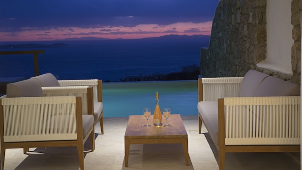 A terrace with wicker furniture and a table with a bottle and glasses, overlooking a body of water and a sunset sky. - Villa Athena (Mykonos)