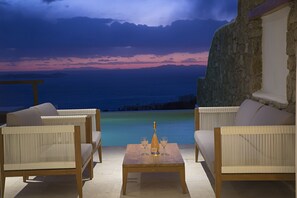 A terrace with wicker furniture and a table with a bottle and glasses, overlooking a body of water and a sunset sky. - Villa Athena (Mykonos)
